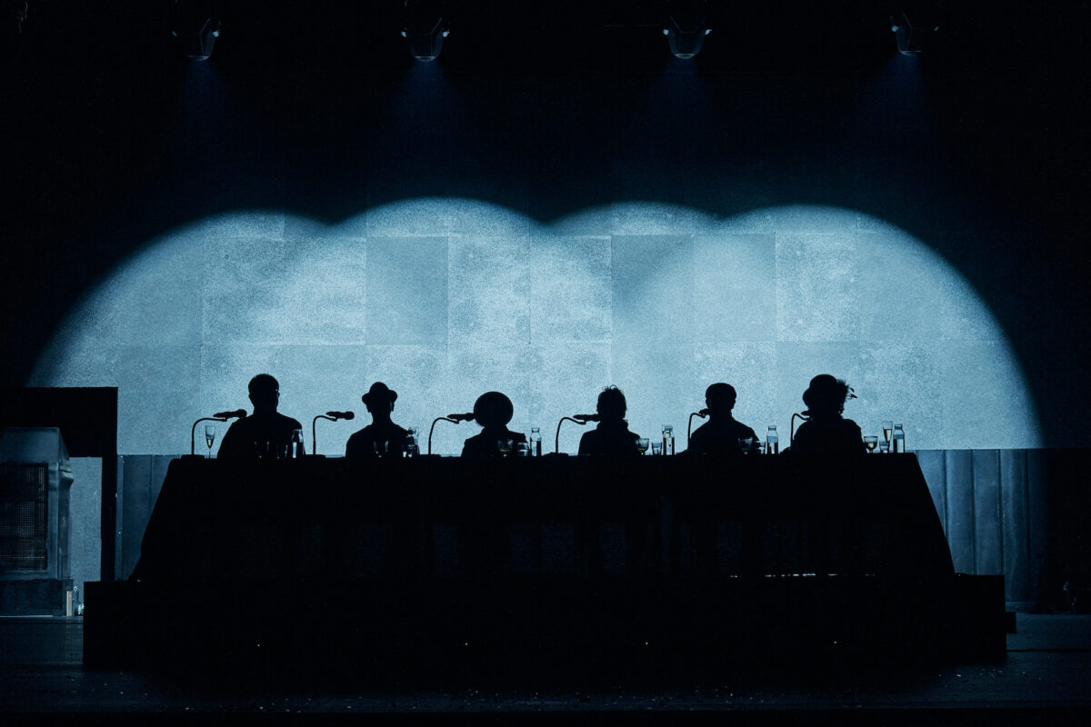 The silhouettes of 6 seniors are dimly lit against a blue stage.