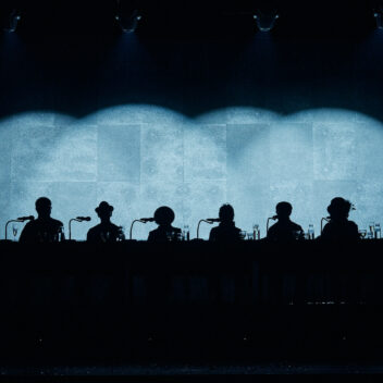 The silhouettes of 6 seniors are dimly lit against a blue stage.